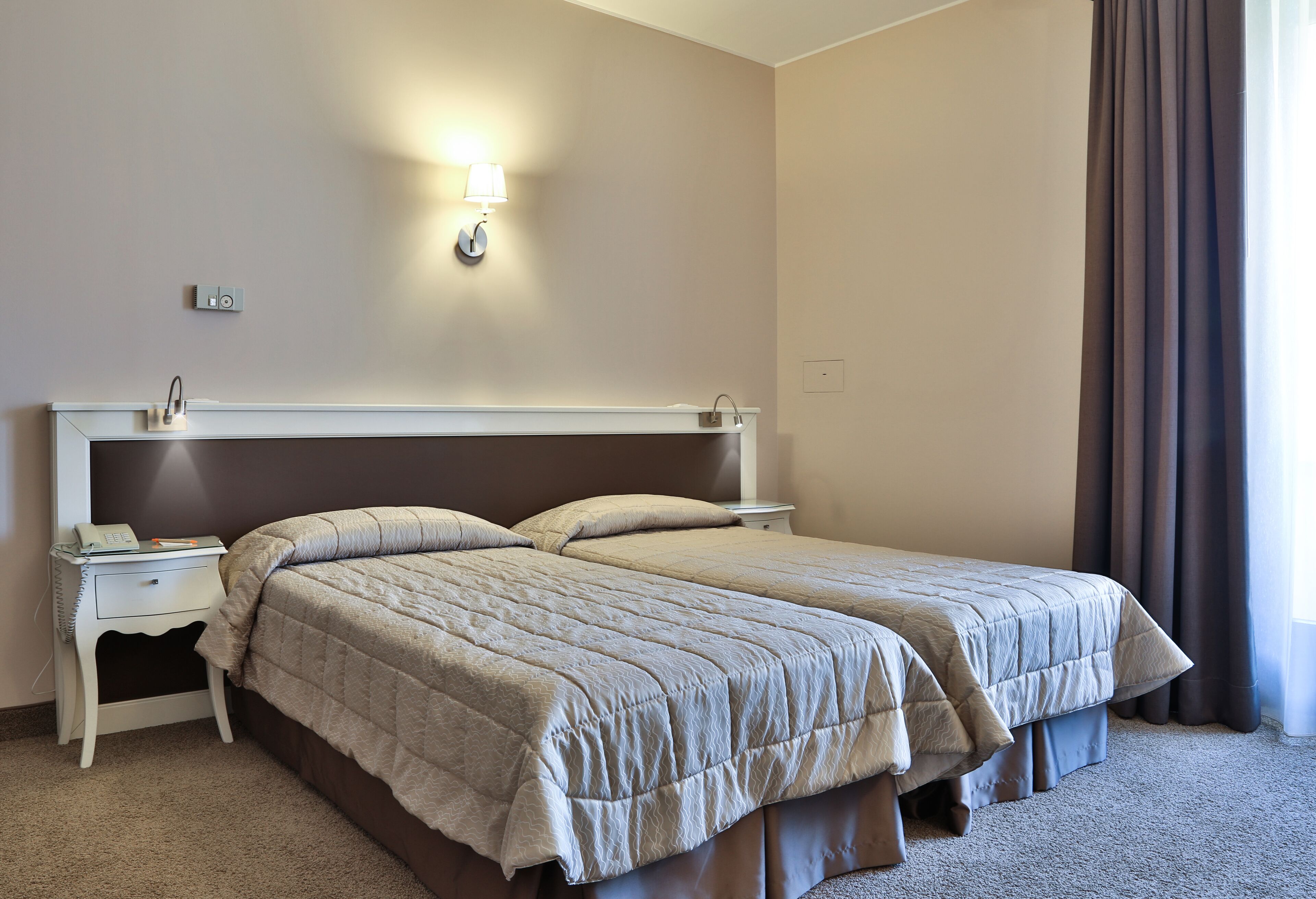 A double bed with a beige quilt, a bedside table with a lamp, and a power outlet on the wall.