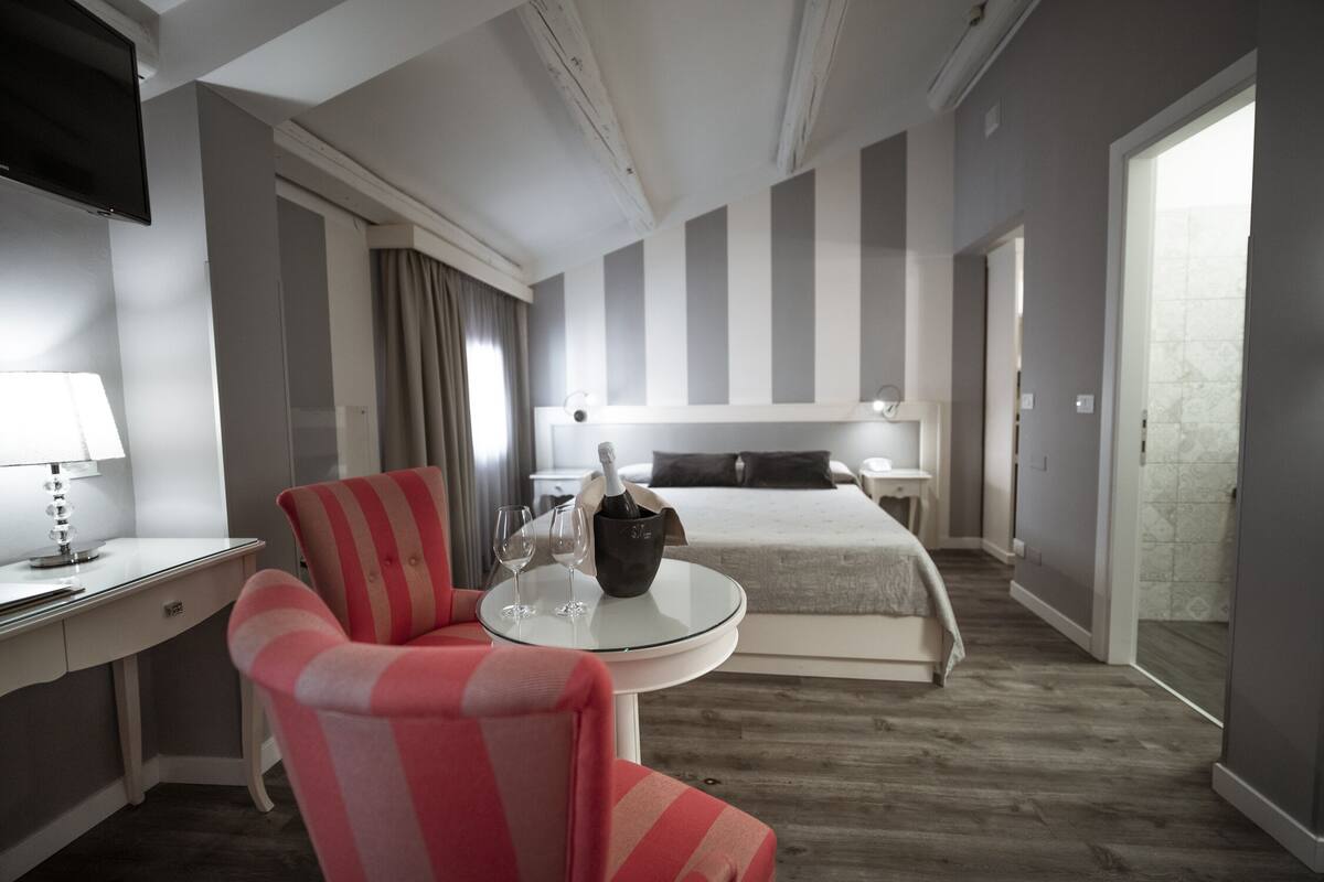 A hotel room with a bed, a desk with a lamp, a chair, and a table with a bottle and glasses.