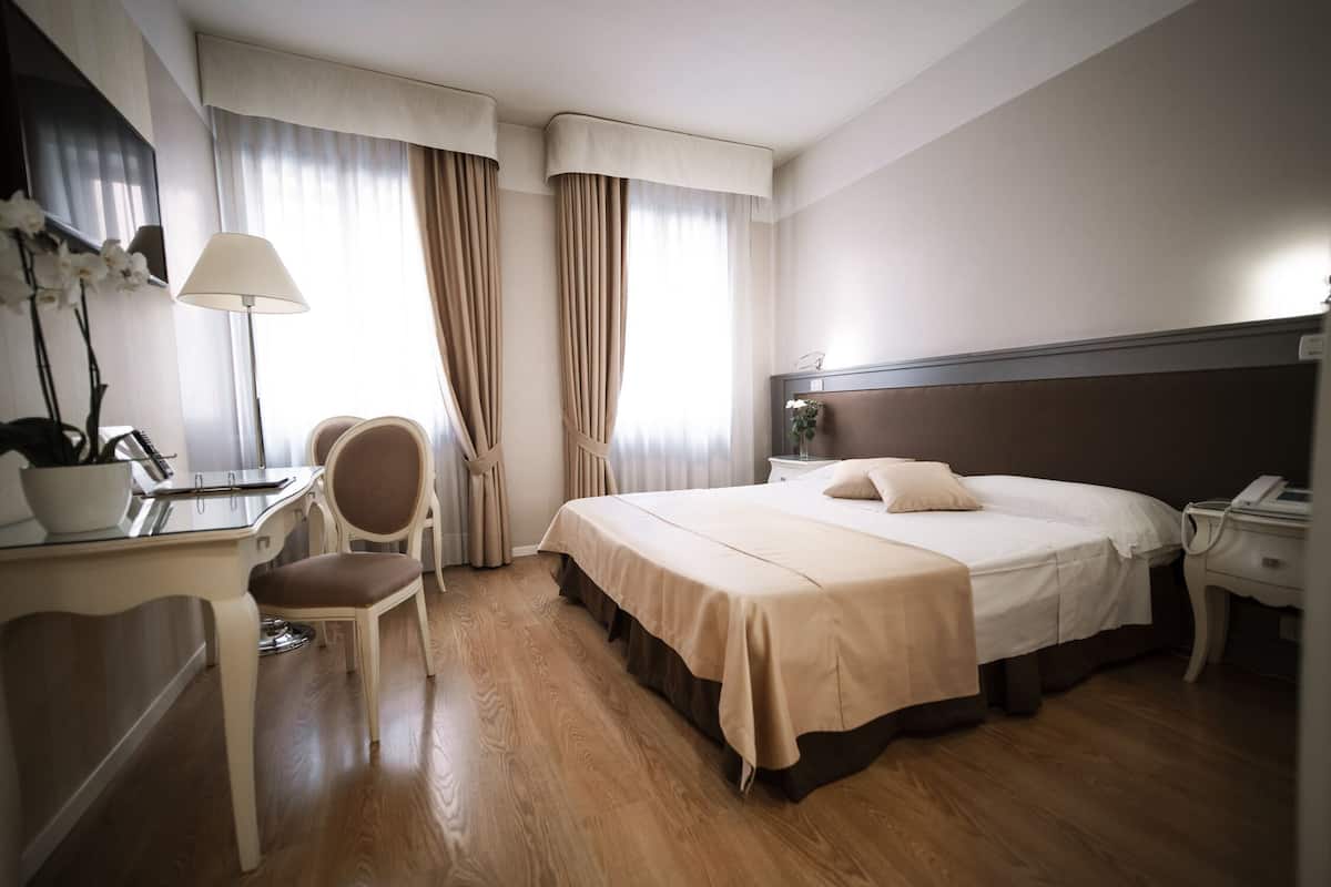A hotel room with a bed, bedside table, lamp, chair, and a desk with a plant.
