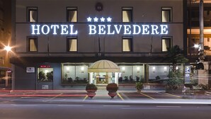 Een hotel genaamd HOTEL BELVEDERE, met een moderne gevel, een groot verlicht bord en een uitnodigende entree met een paraplu.