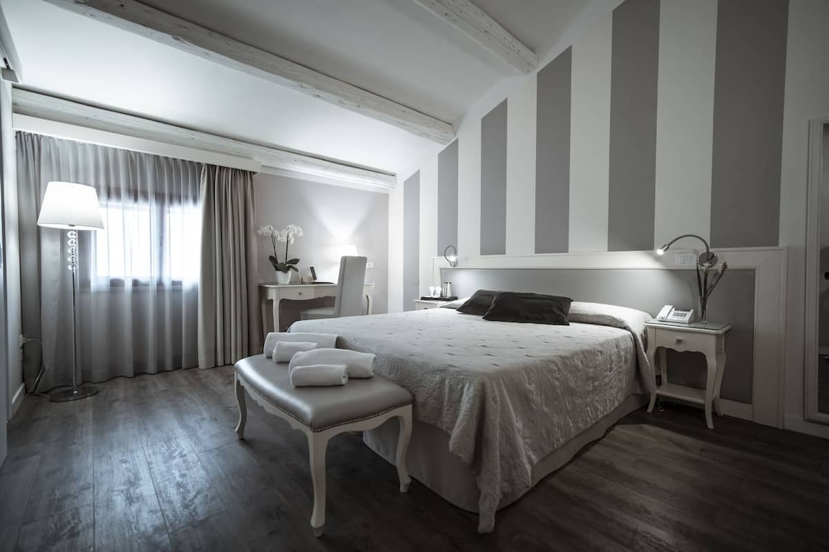 A hotel room with a large bed, a desk, a chair, a bedside table with a lamp, and a window with curtains.