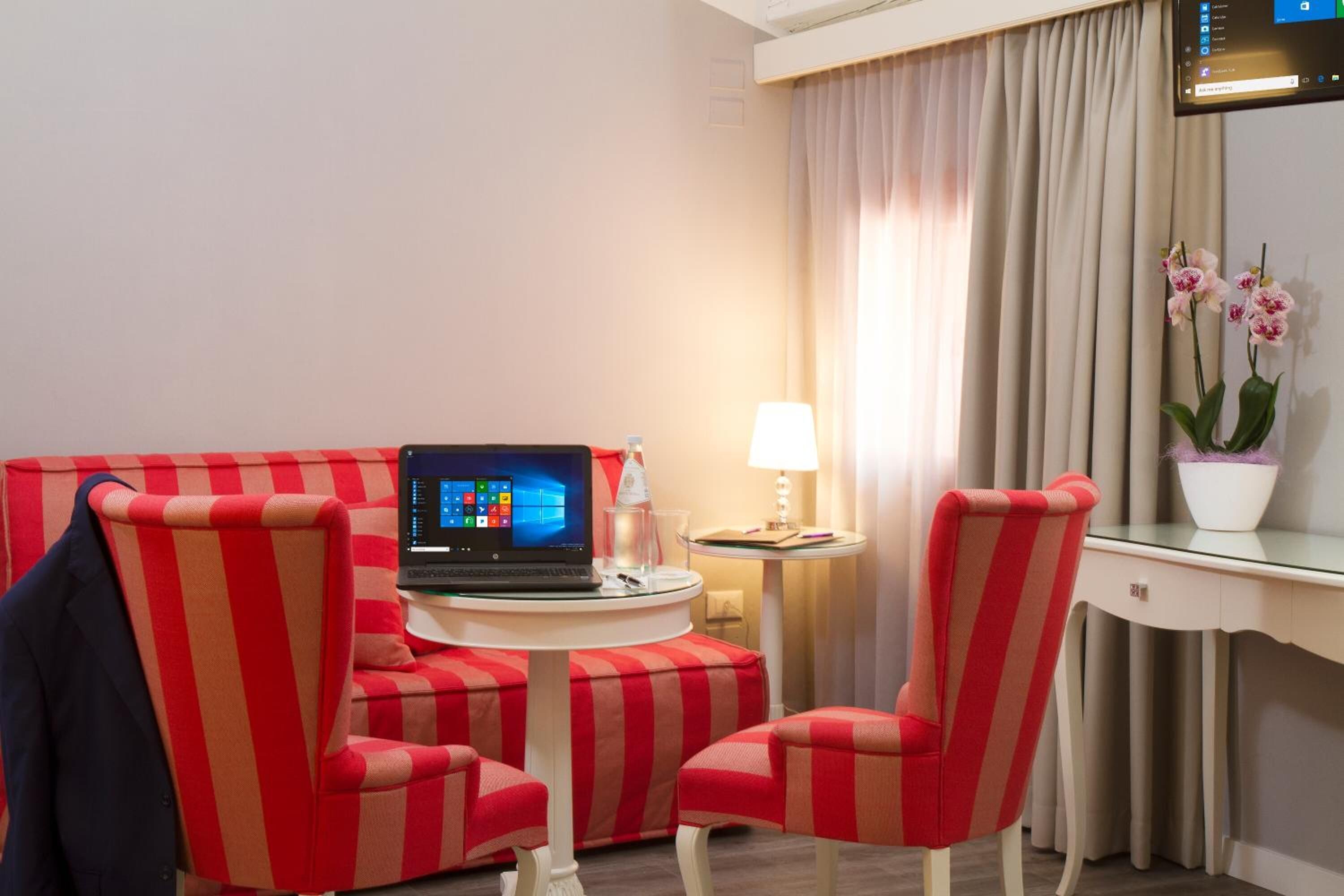 Una habitación de hotel con un sofá a rayas, dos sillones, una computadora portátil sobre una mesita, una lámpara, una planta en maceta y un escritorio con una silla.