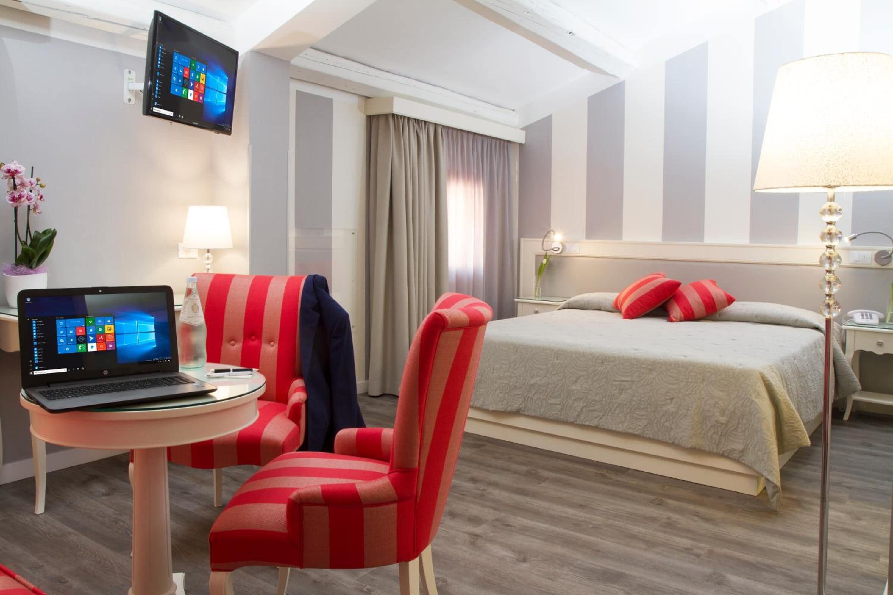 A hotel room with a bed, a desk with a laptop, a striped chair, a lamp, and a TV on the wall.