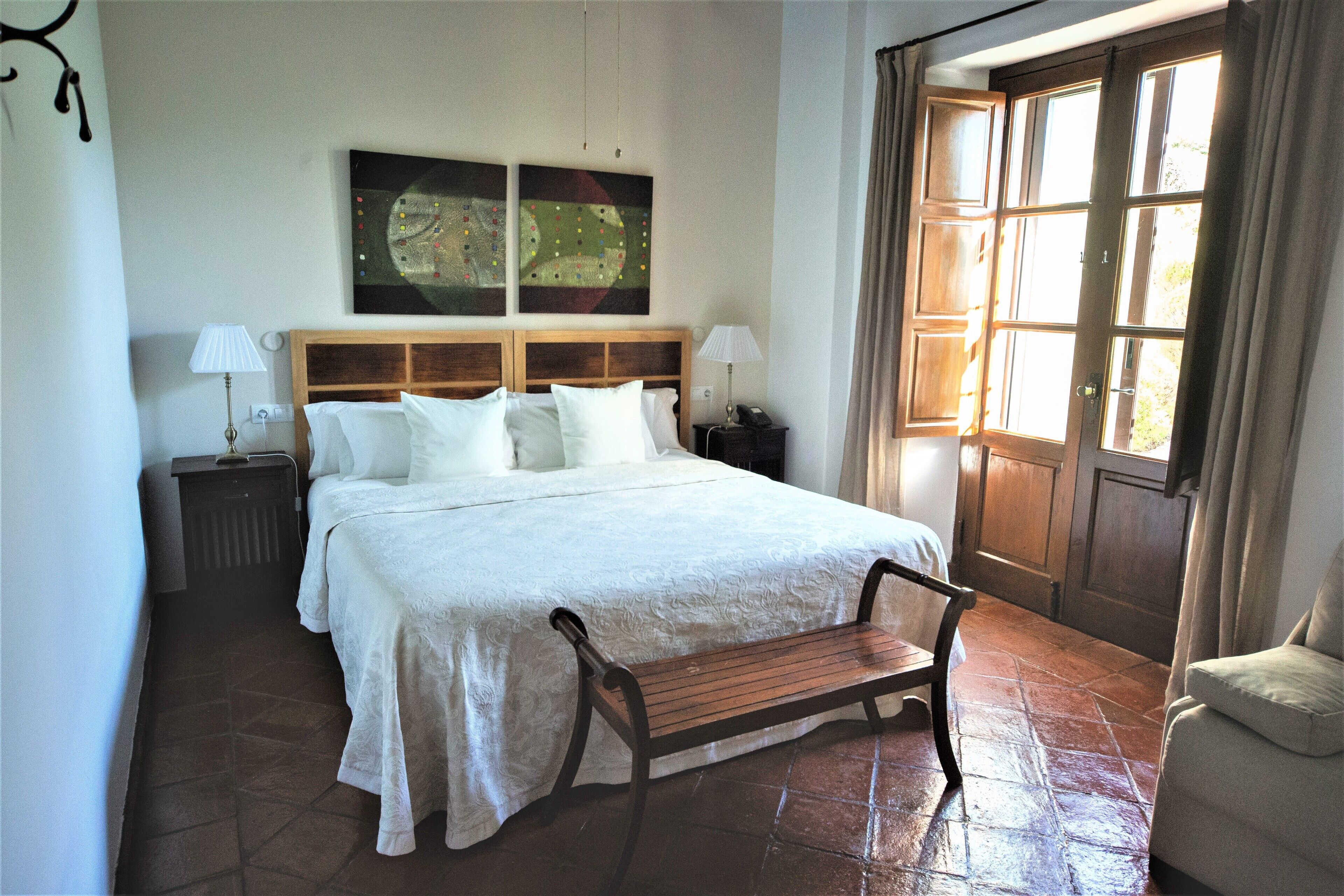 A bedroom with a large bed, two bedside tables, a bench, and a window with curtains.