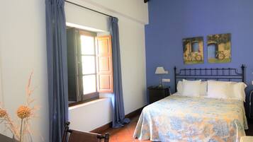 Un dormitorio con cama, mesita de noche, lámpara, ventana con cortinas y dos cuadros en la pared.