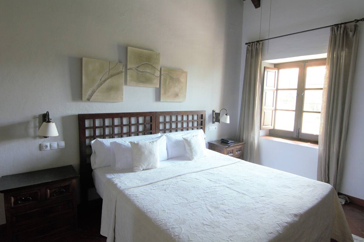 Una cama bien hecha con sábanas blancas, cabecera de madera y tres obras de arte colgadas en la pared.