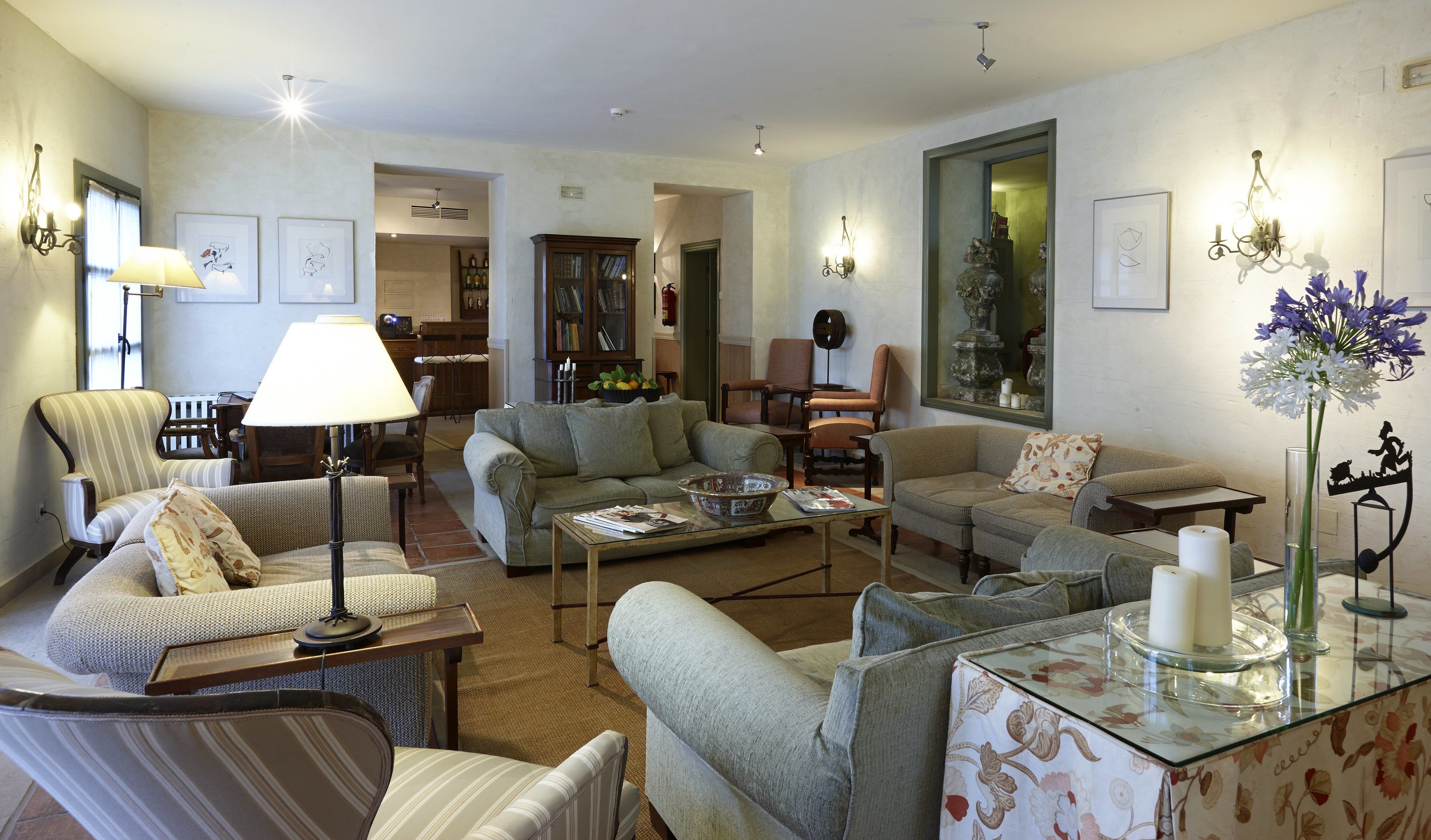 A cozy living room with a sofa, armchairs, a coffee table, and a lamp. There is a bookshelf and a window with curtains.