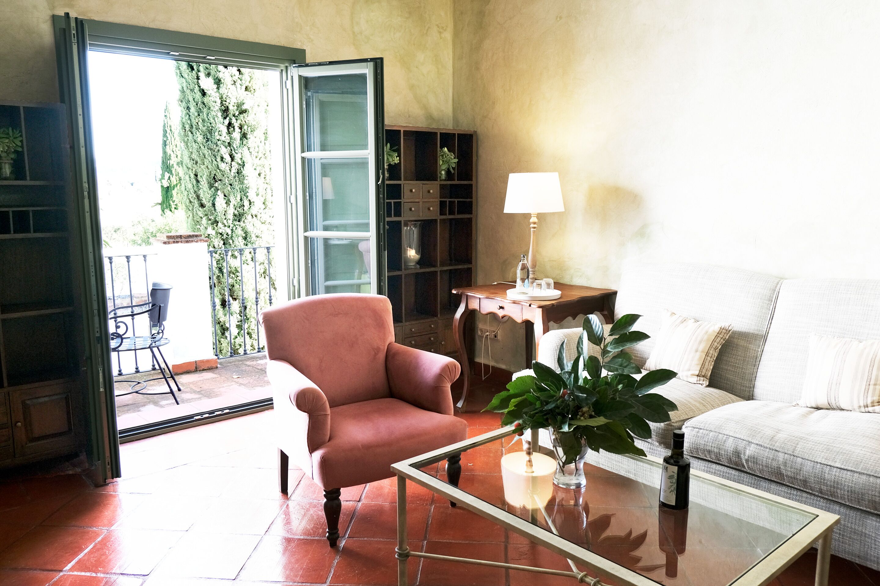 Una sala de estar con una mesa de centro de vidrio, un sillón rosa, un sofá blanco, un escritorio de madera con una lámpara y una estantería.