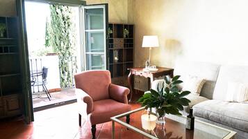 Una sala de estar con una mesa de centro de vidrio, un sillón rosa, un sofá blanco, un escritorio de madera con una lámpara y una estantería.