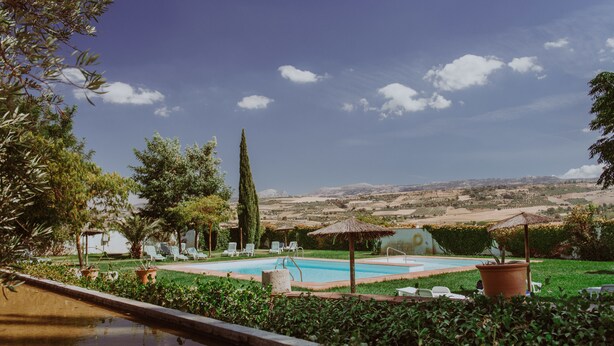A pool area with lounge chairs and umbrellas, surrounded by greenery and a view of distant hills. - Hotel Boutique Molino del Arco (Ronda)