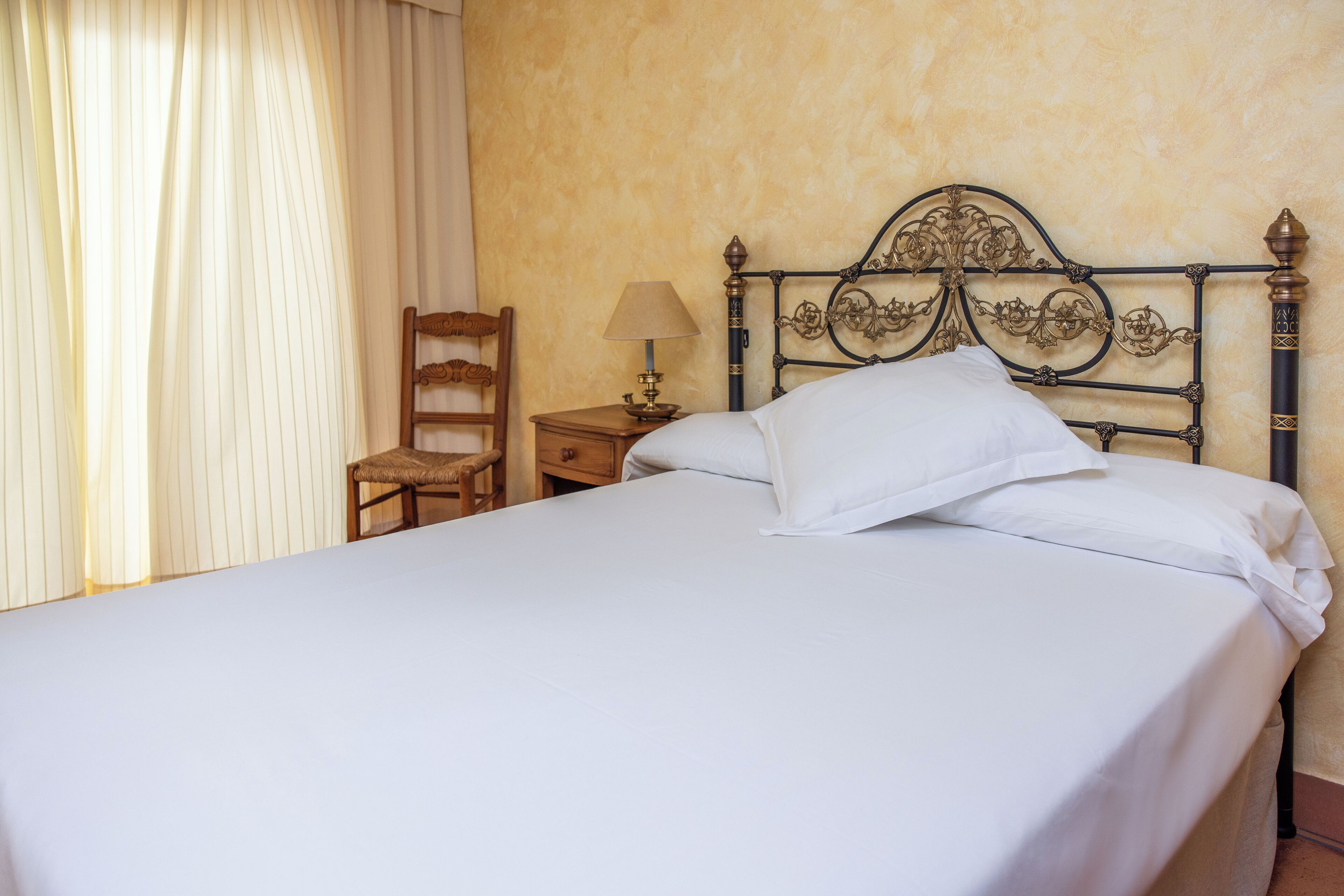 A neatly made bed with white linens, a wooden chair, a bedside table with a lamp, and a decorative metal headboard.
