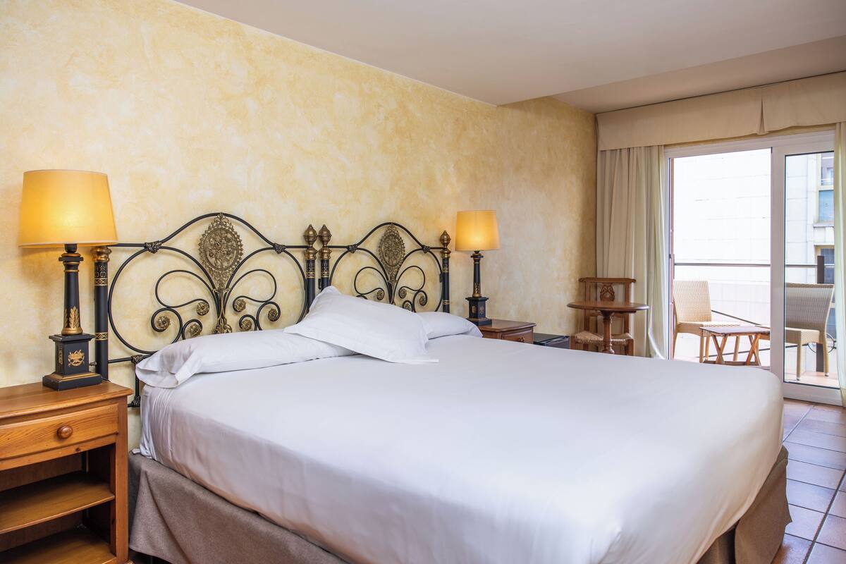 Una cama bien hecha con ropa de cama blanca, dos lámparas de noche, una mesita de madera y una silla junto a una puerta corrediza de vidrio.