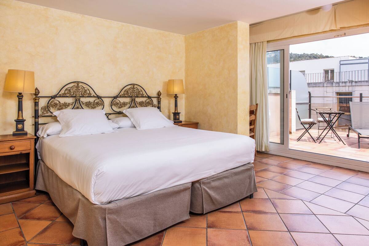 Habitación de hotel con una cama grande, dos mesitas de noche con lámparas, vista al balcón exterior a través de una puerta corrediza y piso de baldosas.