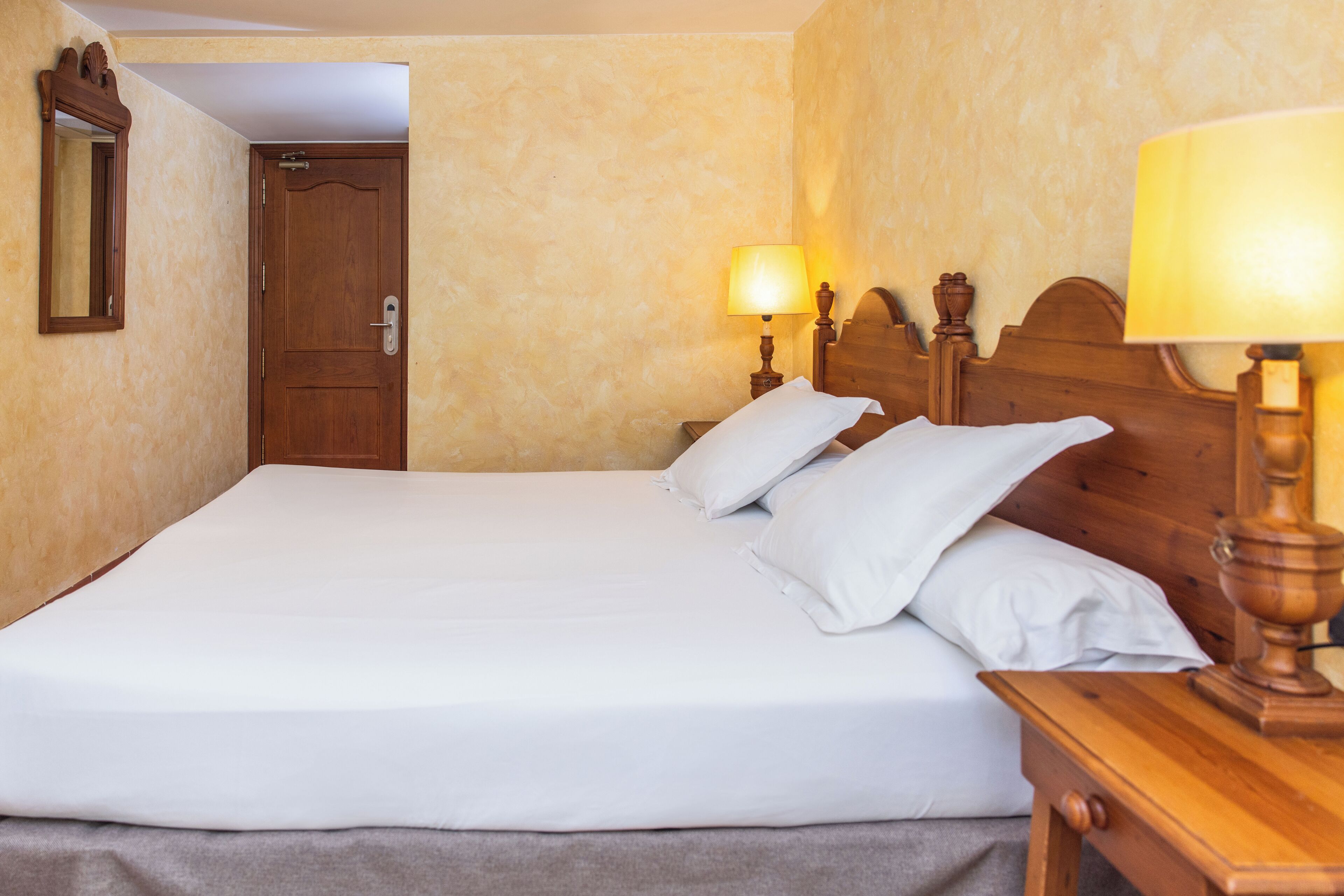 A neatly made bed with white linens and pillows, a wooden nightstand with a lamp, and a wooden door in the background.