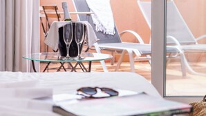 Een hotelkamer met een glazen tafel waarop twee champagneglazen, een zonnebril en een boek staan. Er zijn twee fauteuils met witte kussens en een handdoek is over een ervan gedrapeerd. De kamer heeft een groot raam met uitzicht op het buitengebied.