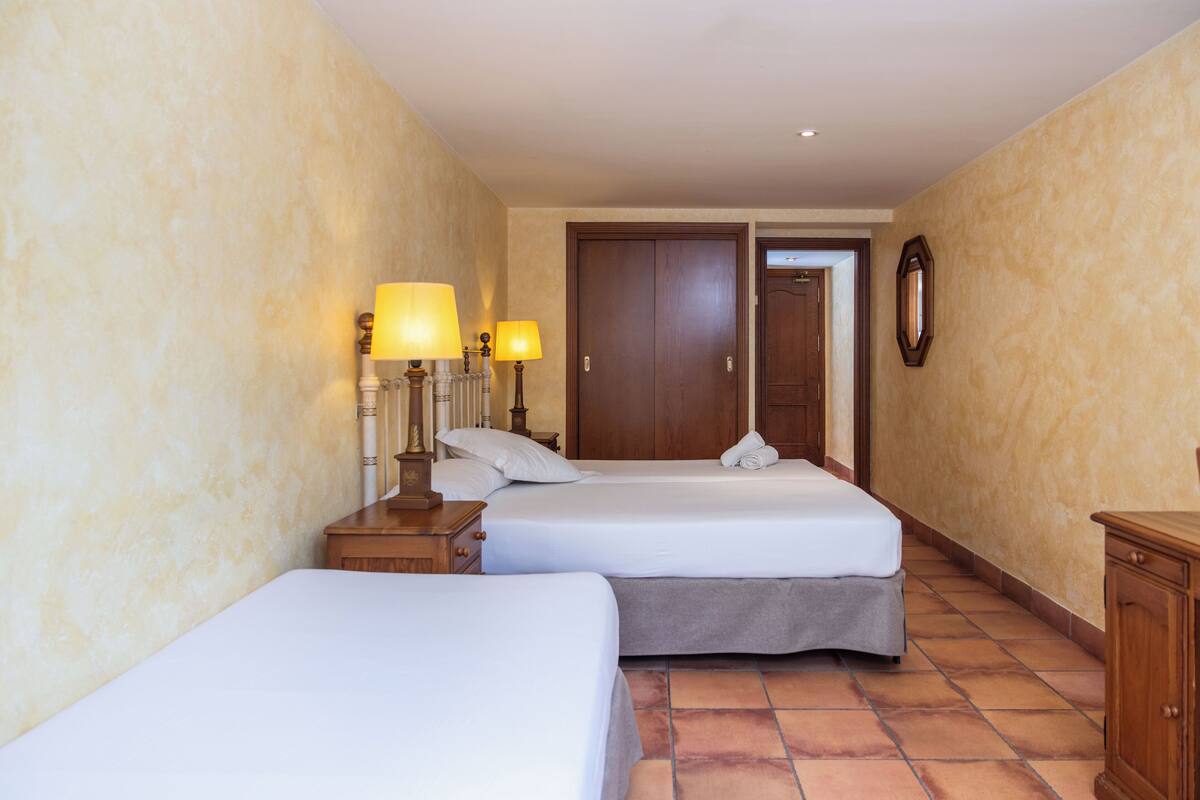 Habitación de hotel con dos camas, una mesita de noche con lámparas, un armario de madera y un espejo en la pared.