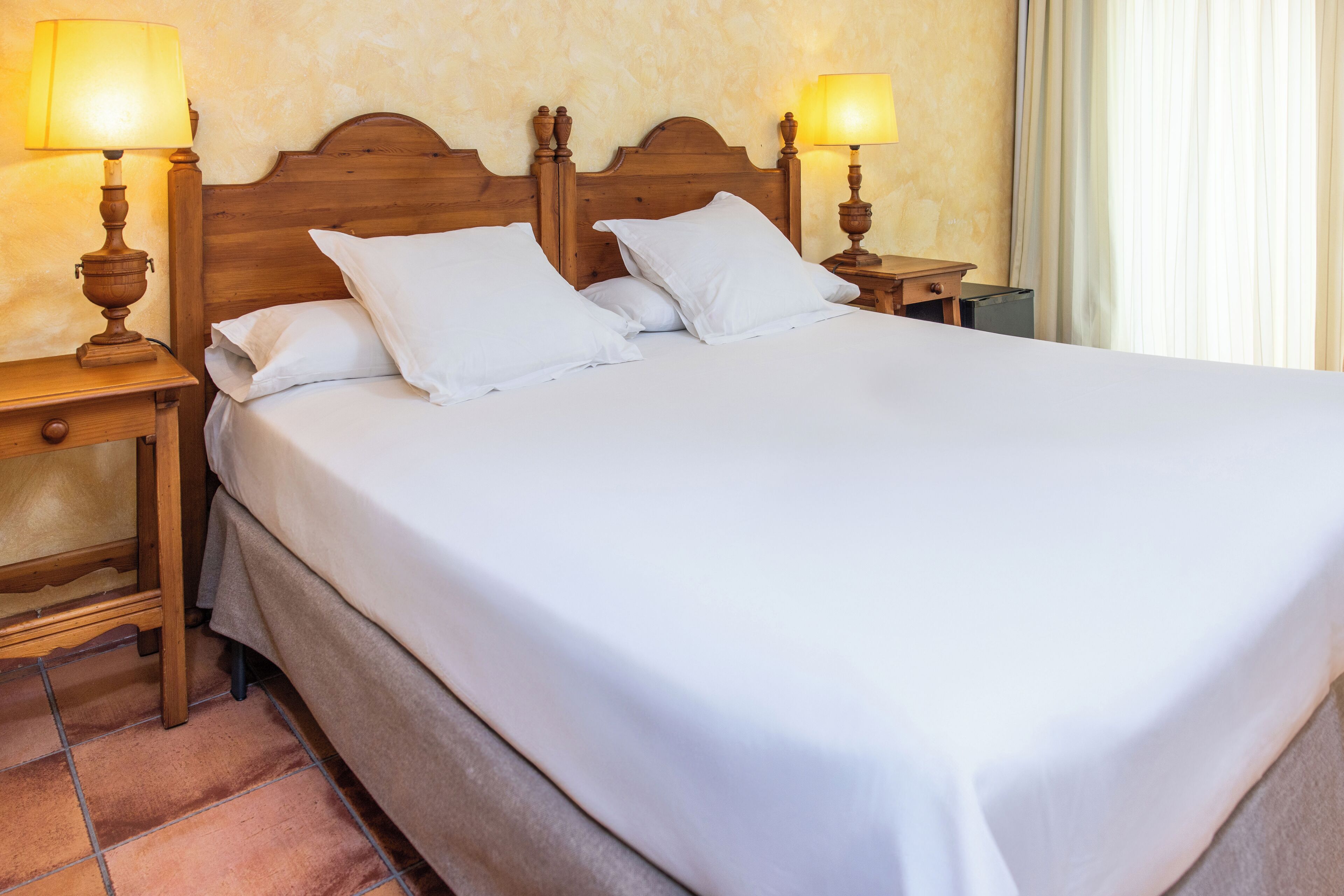 Una cama bien hecha con sábanas blancas y dos lámparas de noche sobre mesitas de madera.