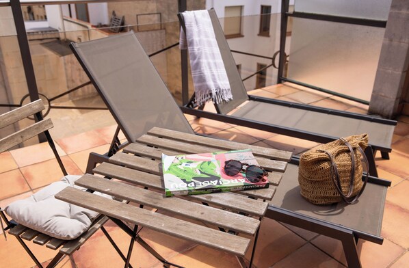 A sun lounger with a magazine, sunglasses, and a woven bag on a terrace with tiled flooring.