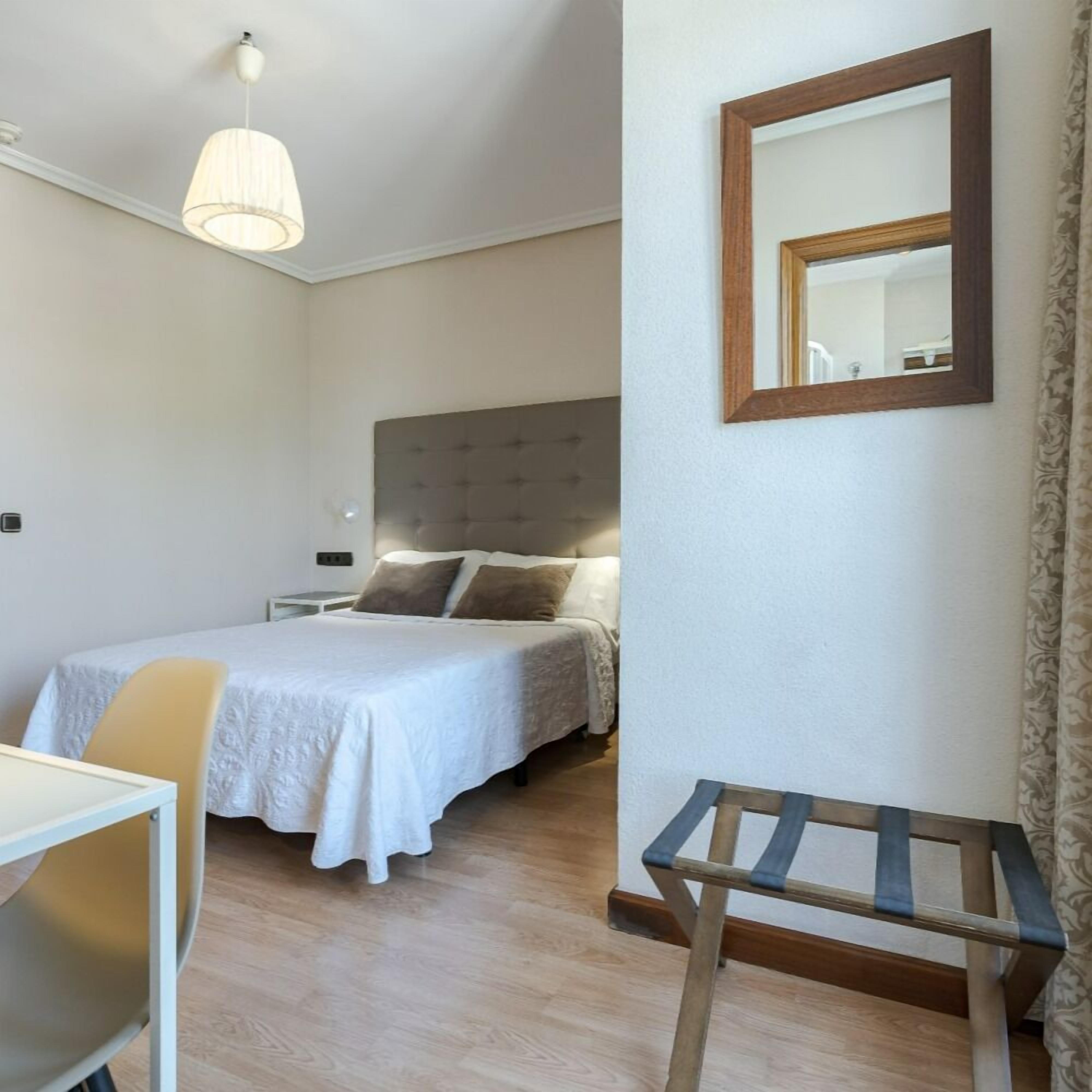 A hotel room with a bed, a bedside table, a chair, a hanging light fixture, and a mirror on the wall.
