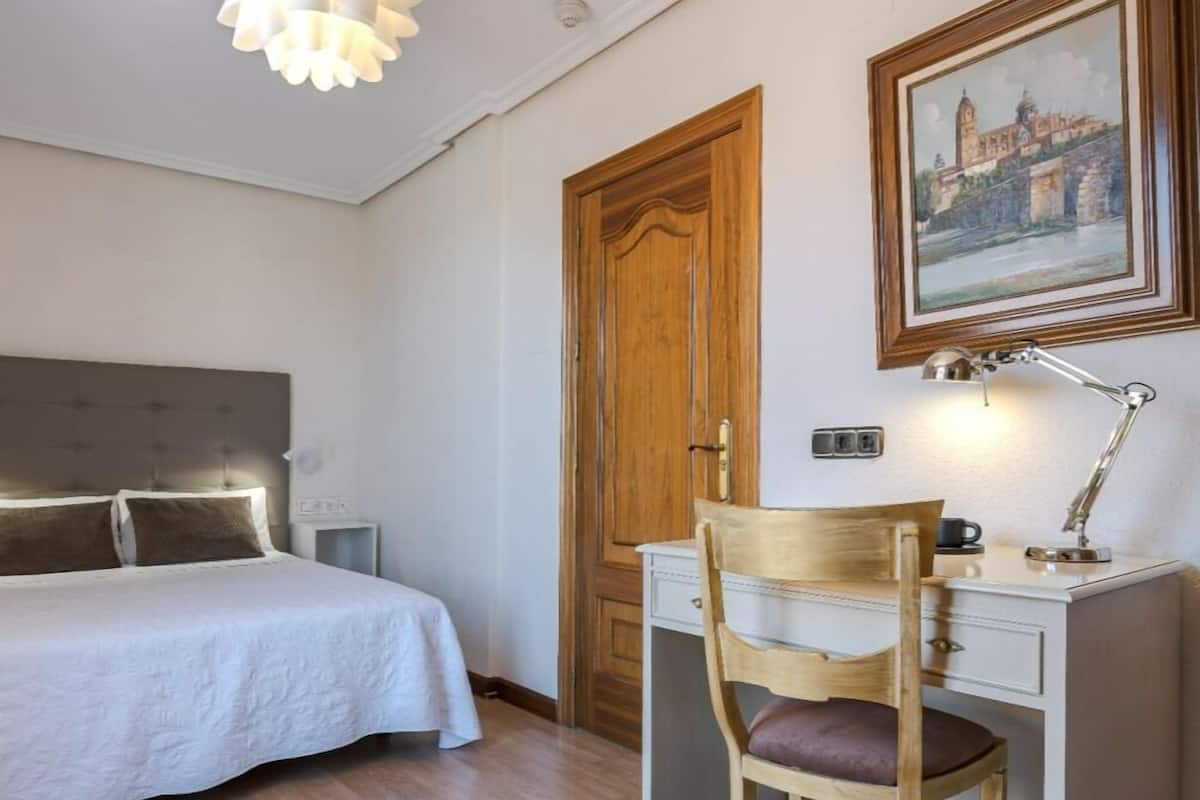 A bedroom with a bed, a desk, a chair, a desk lamp, and a painting on the wall.