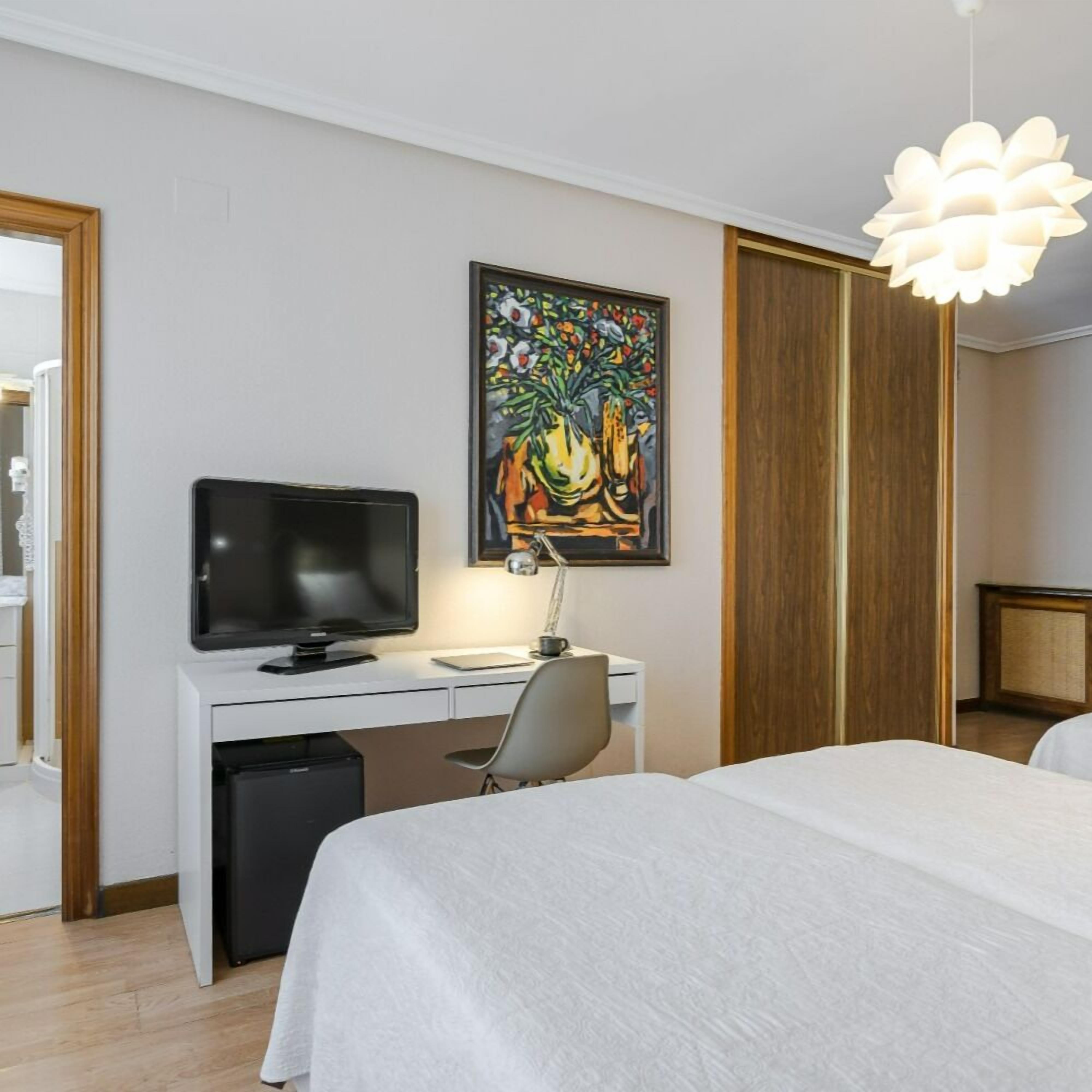 Une chambre d’hôtel avec un lit, un bureau équipé d’un ordinateur, une télévision, un tableau au mur et un luminaire au plafond au design original.
