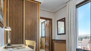 A hotel room with a wooden wardrobe, a desk with a lamp, a chair, a small table, a large window with a view, and a mirror on the wall. - Hotel Salamanca Rio (Salamanca)