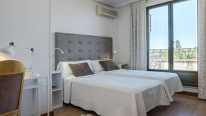 A bedroom with a grey tufted headboard, two beds with white linens, a desk with a lamp, and a large window with a view of greenery. - Hotel Salamanca Rio (Salamanca)