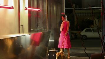 Una persona vestida de rosa está de pie en un vestíbulo moderno, con un perro, una maleta y un vidrio que da al exterior.