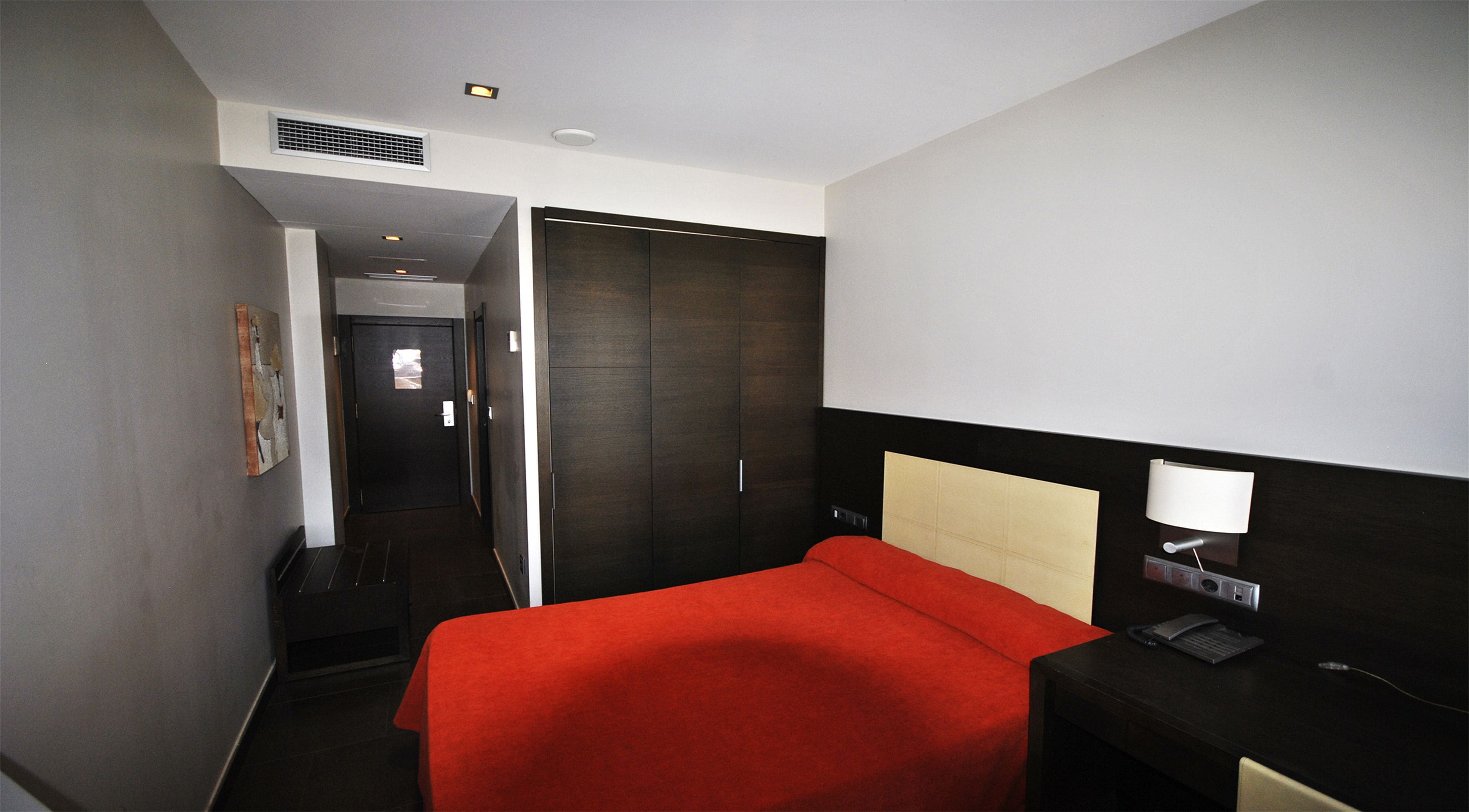 A hotel room with a bed, a desk with a lamp, a wardrobe, and a door leading to a hallway.