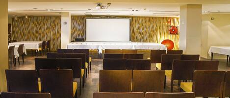 Un salón de conferencias con filas de sillas, una gran mesa con una pantalla para proyector y un extintor de incendios en la pared.