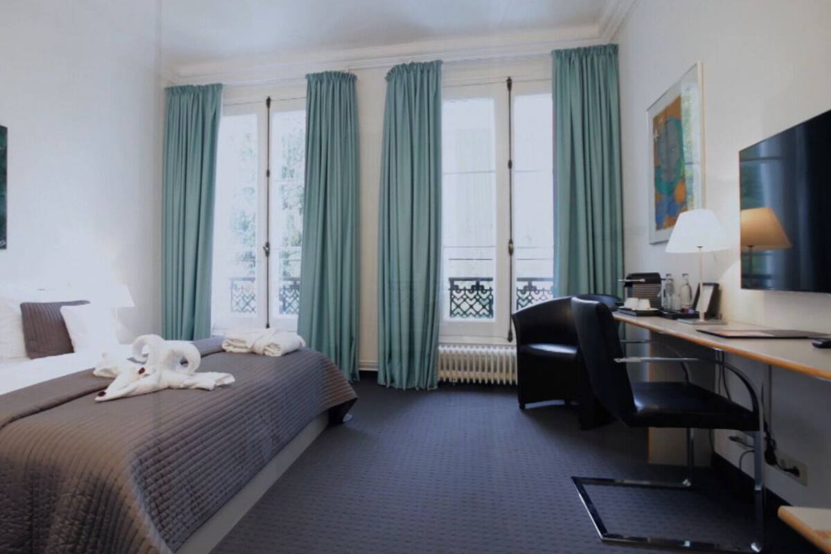 A hotel room with a bed, desk, chair, and large windows with curtains.