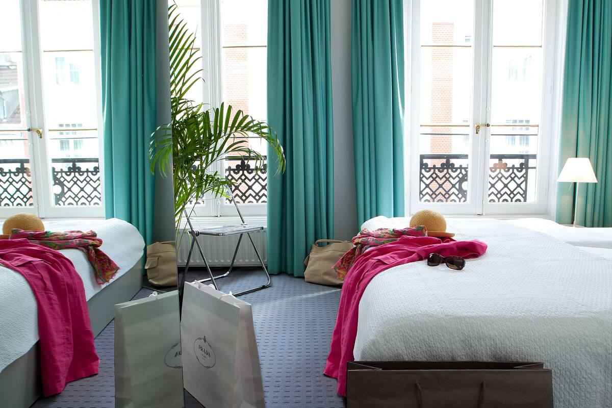 A hotel room with two beds, teal curtains, a potted plant, and shopping bags.