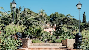 A garden pathway leading to a building with a red facade, surrounded by palm trees and a stone wall. - Hotel Villa Retiro (Xerta)