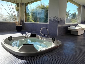 A modern spa area with a glass-enclosed hot tub, a seating area, and a view of greenery through large windows. - Hotel Villa Retiro (Xerta)