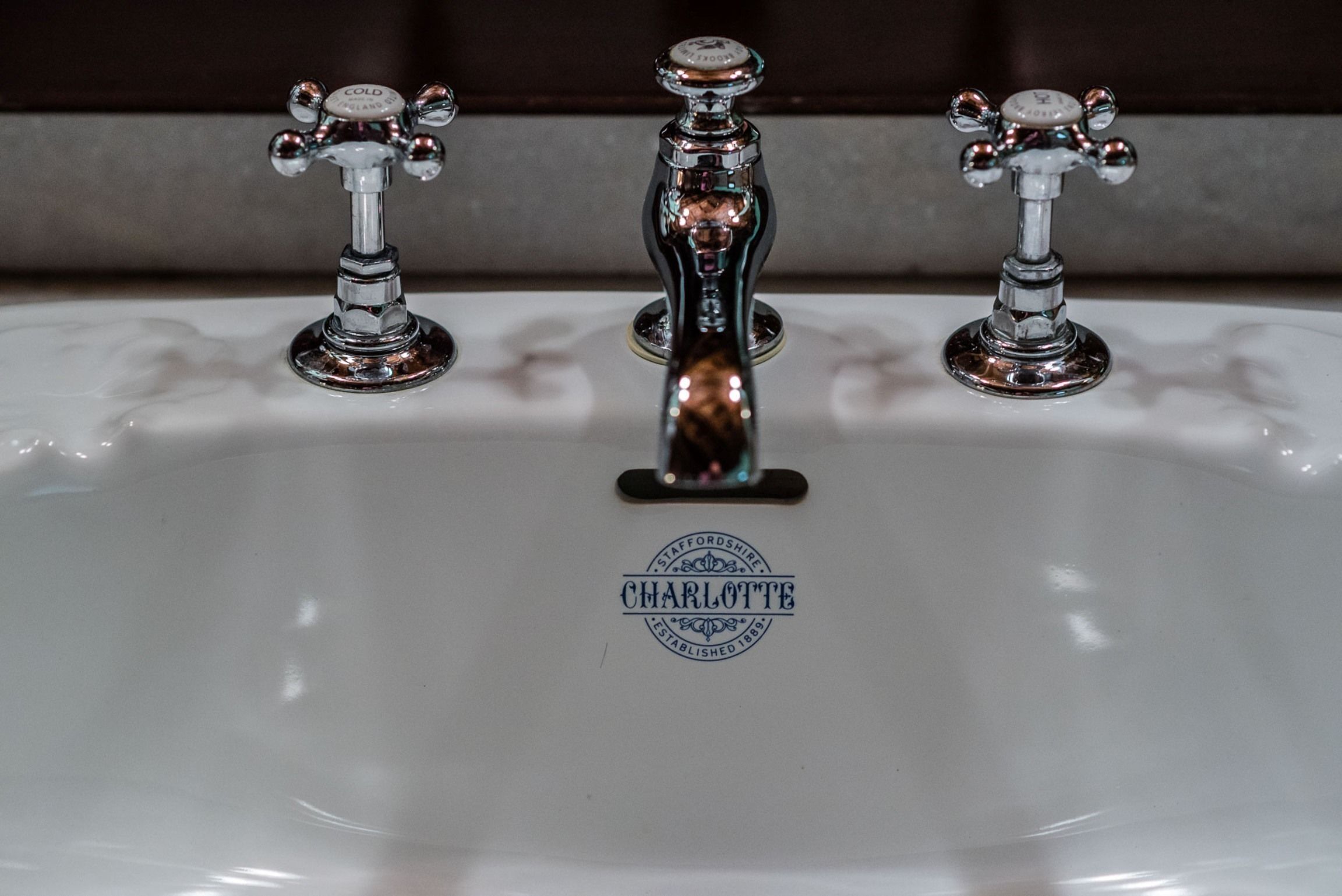 A bathroom sink with three faucets, one central and two side. The central faucet has a label that reads "CHARLOTTE" and "STAFFORDSHIRE ESTABLISHED 1899."