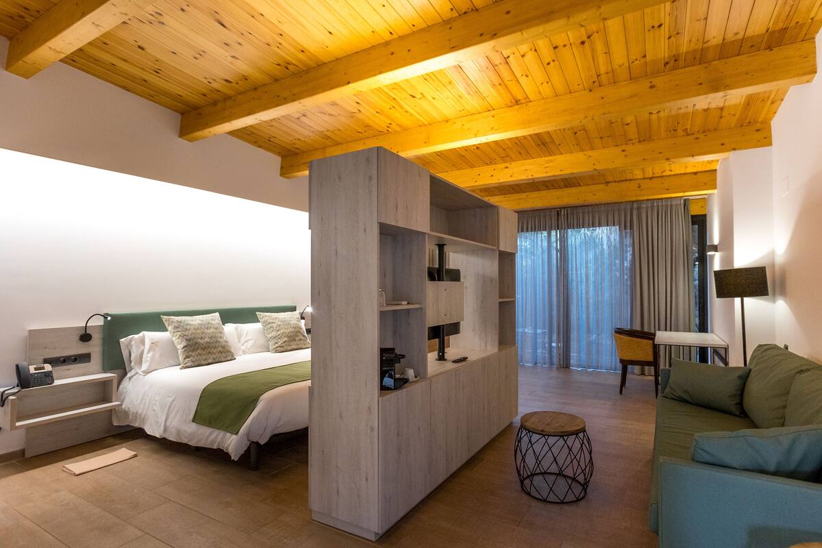Una habitación de hotel moderna con techo de madera, cama con cabecera verde, sofá, una pequeña mesa redonda y un mueble para televisión.