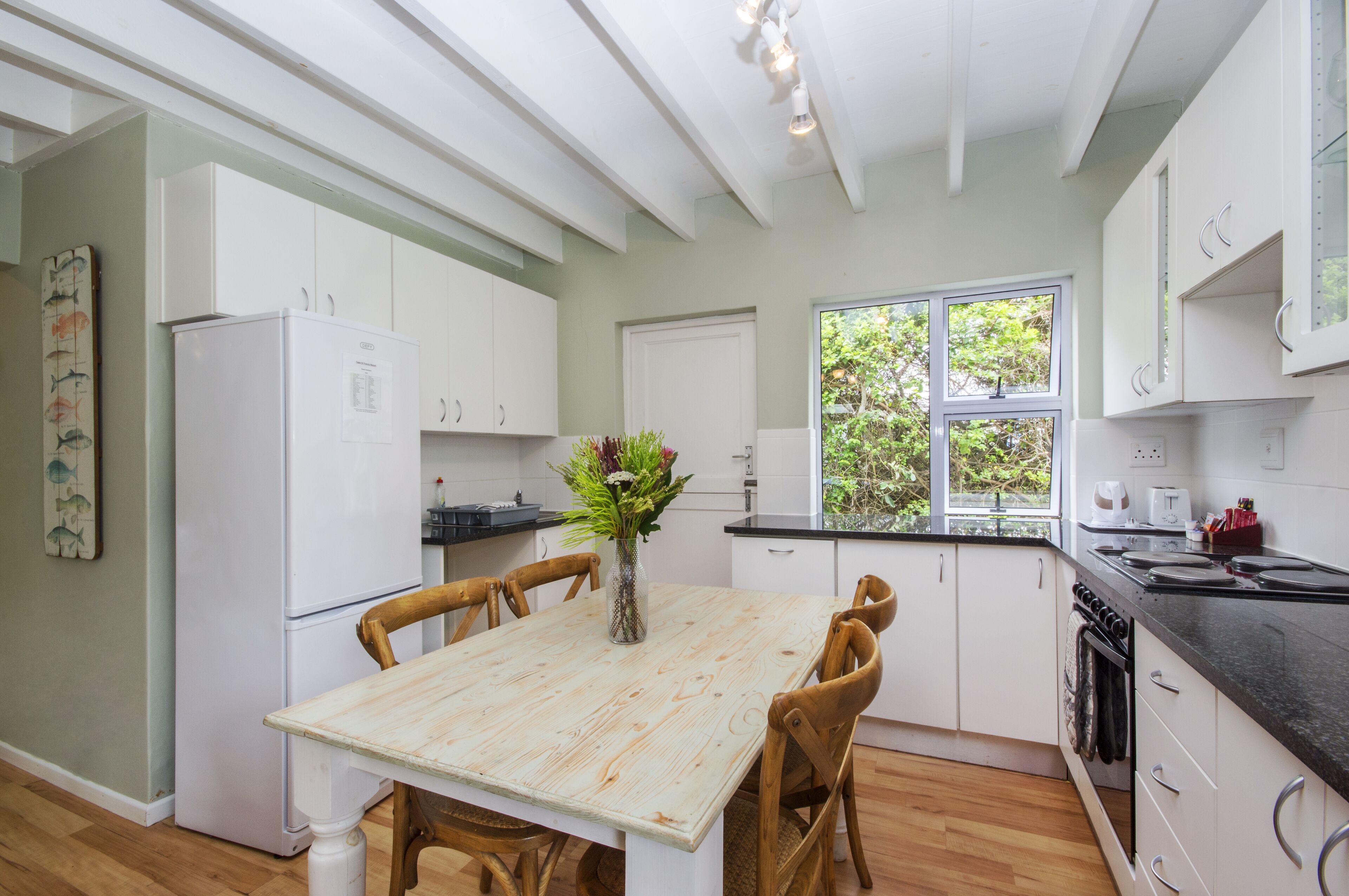 A modern kitchen with white cabinets, a wooden dining table, and a window with a view of greenery.