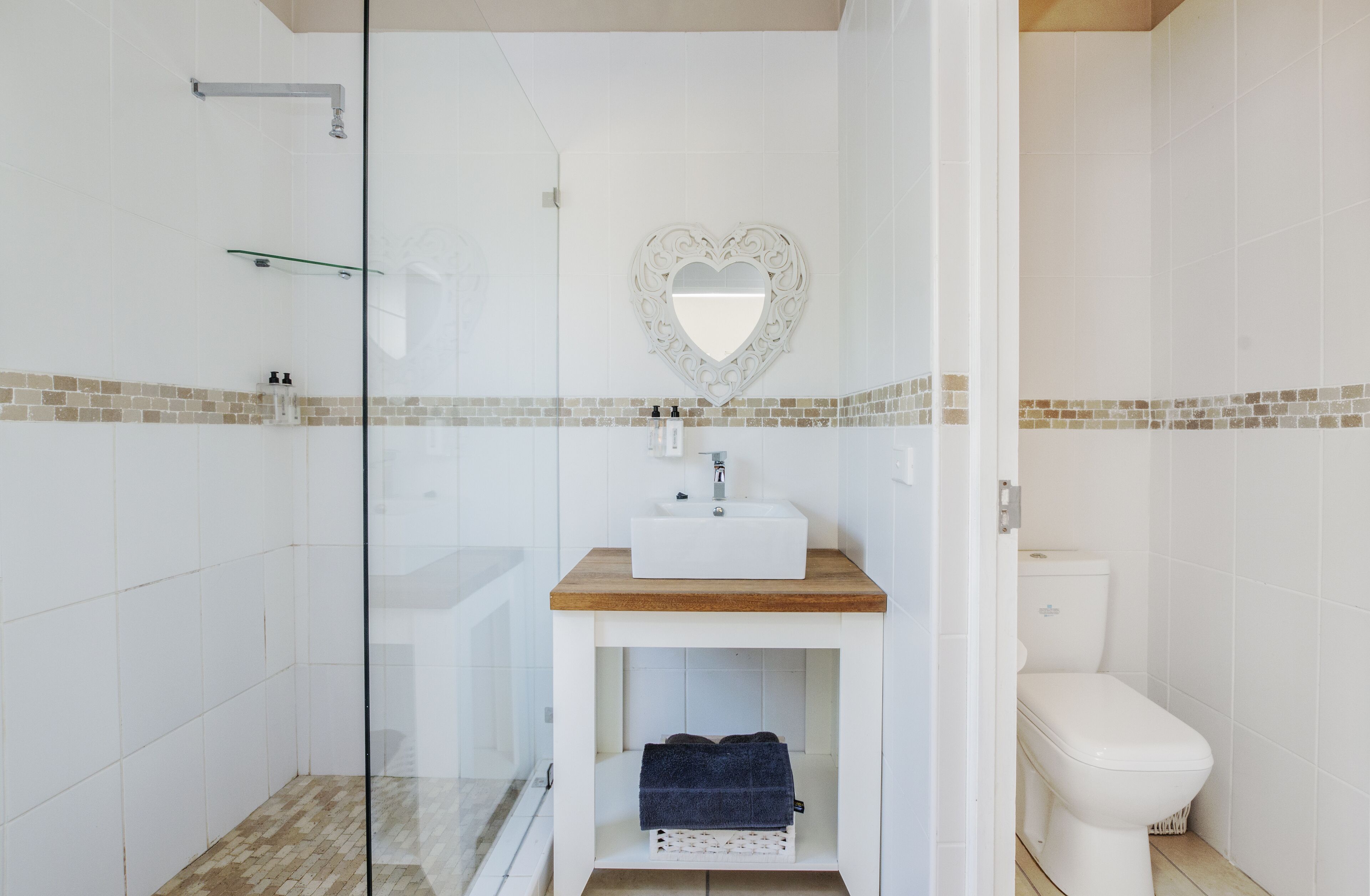 A bathroom with a glass shower enclosure, a white sink with a wooden countertop, a heart-shaped mirror, and a toilet.