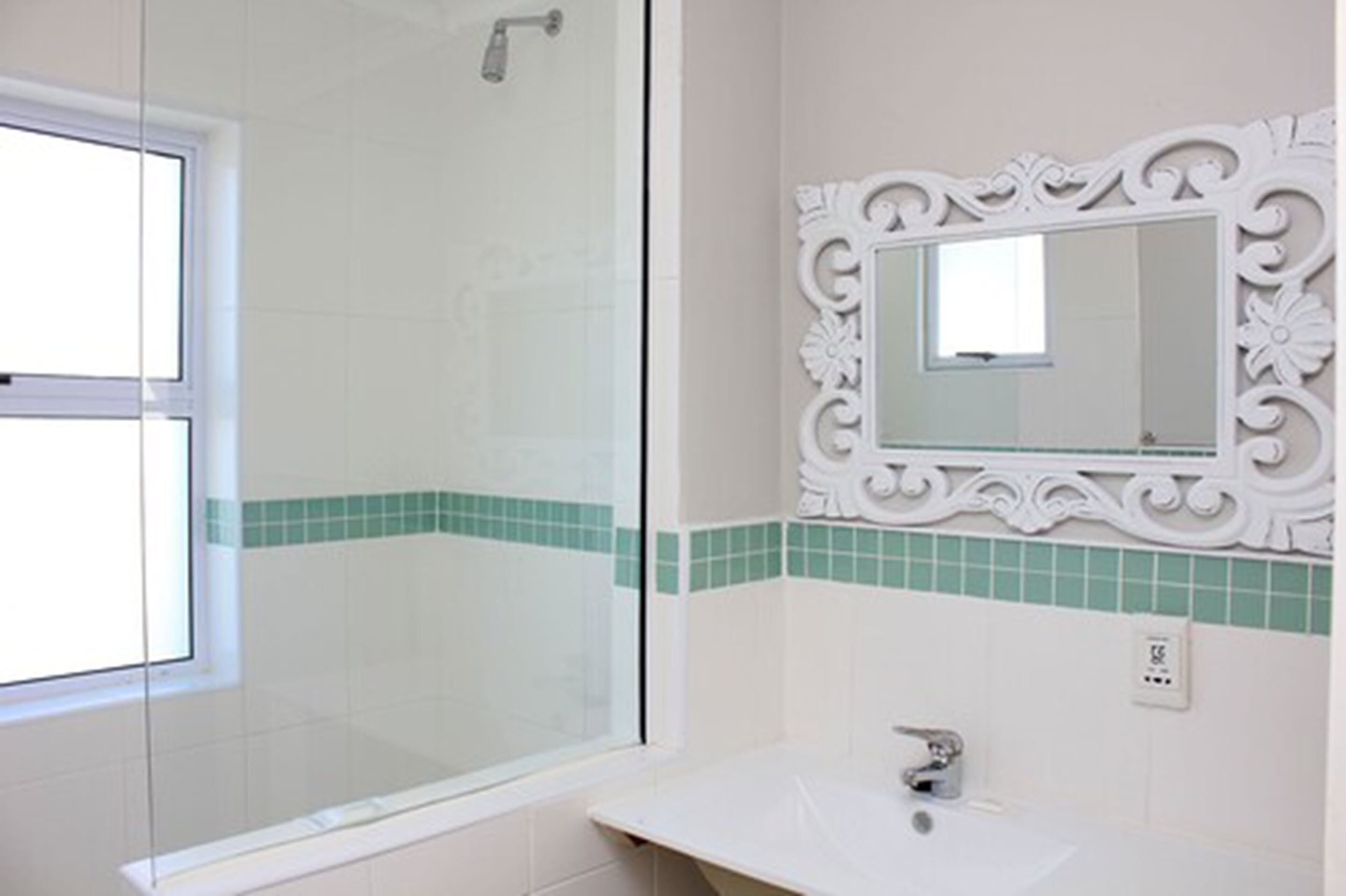 A bathroom with a glass shower enclosure, a white sink, and a decorative mirror with a green-tiled backsplash.