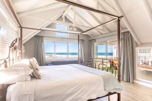 A spacious bedroom with a four-poster bed, large windows, and a balcony overlooking the ocean.