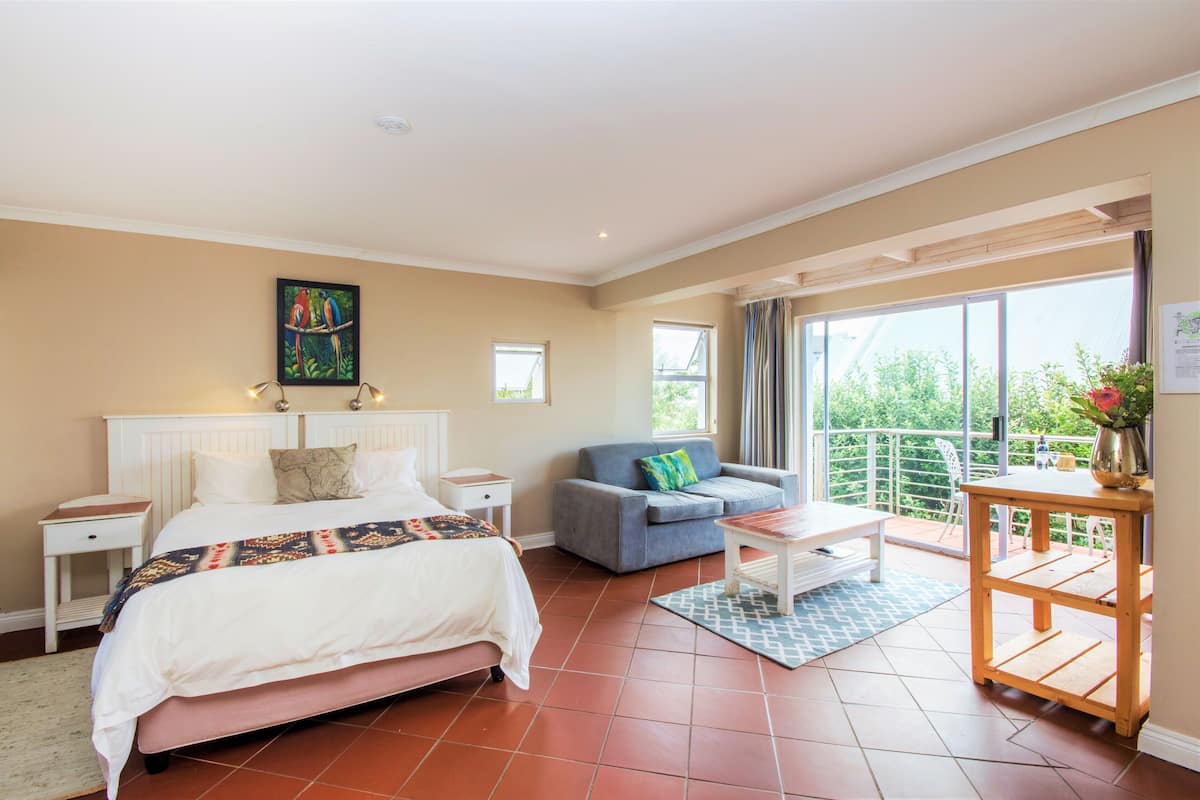 Een slaapkamer met een bed, een grijze bank, een kleine tafel en een balkon met uitzicht op groen.