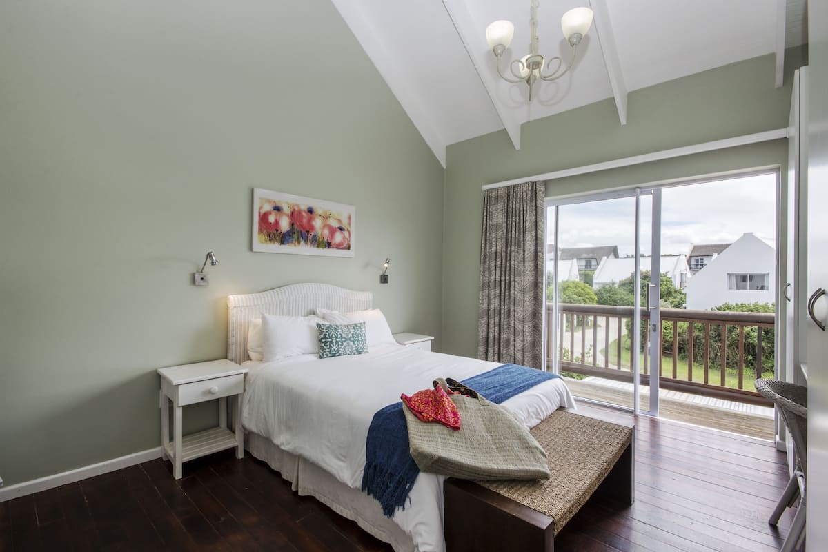 Een slaapkamer met een bed, nachtkastje, schilderij en een balkon met uitzicht op huizen.