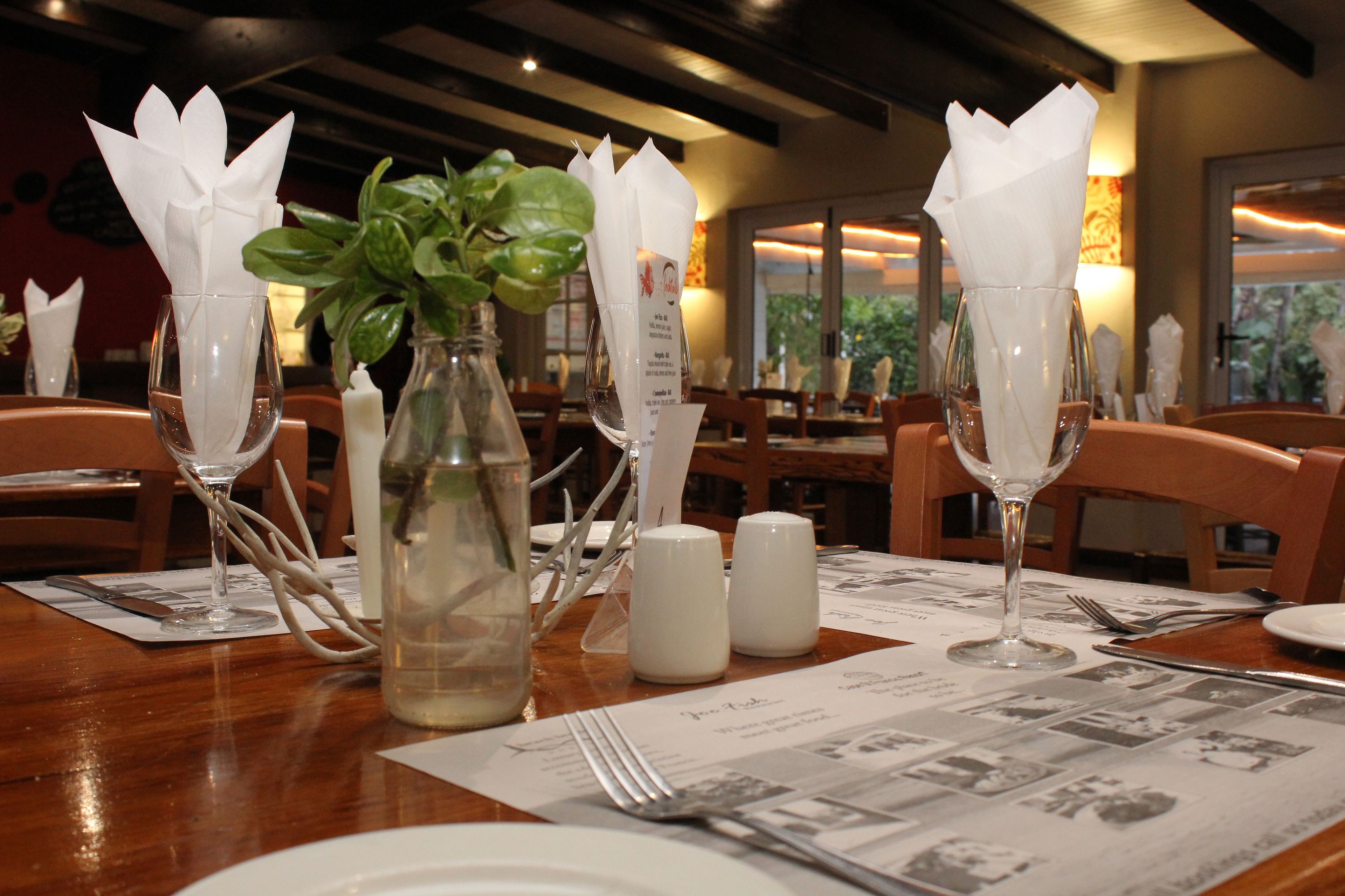 Une table à manger dressée avec des serviettes blanches, des salières et des poivrières, un vase en verre contenant des feuilles vertes et un menu.