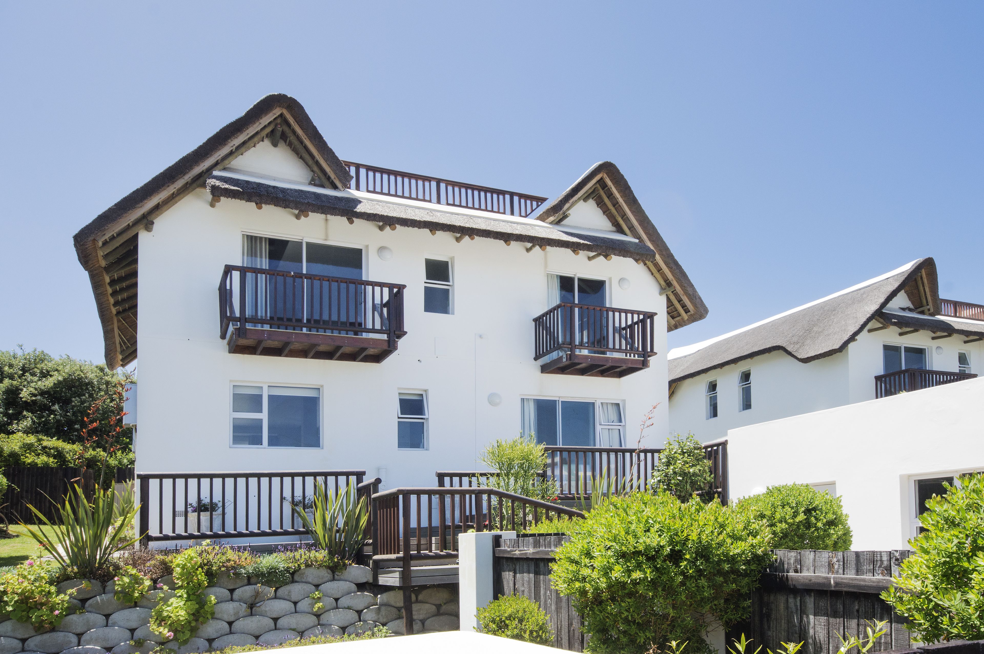 A two-story residential building with a white exterior, wooden balconies, and a stone retaining wall in the foreground.