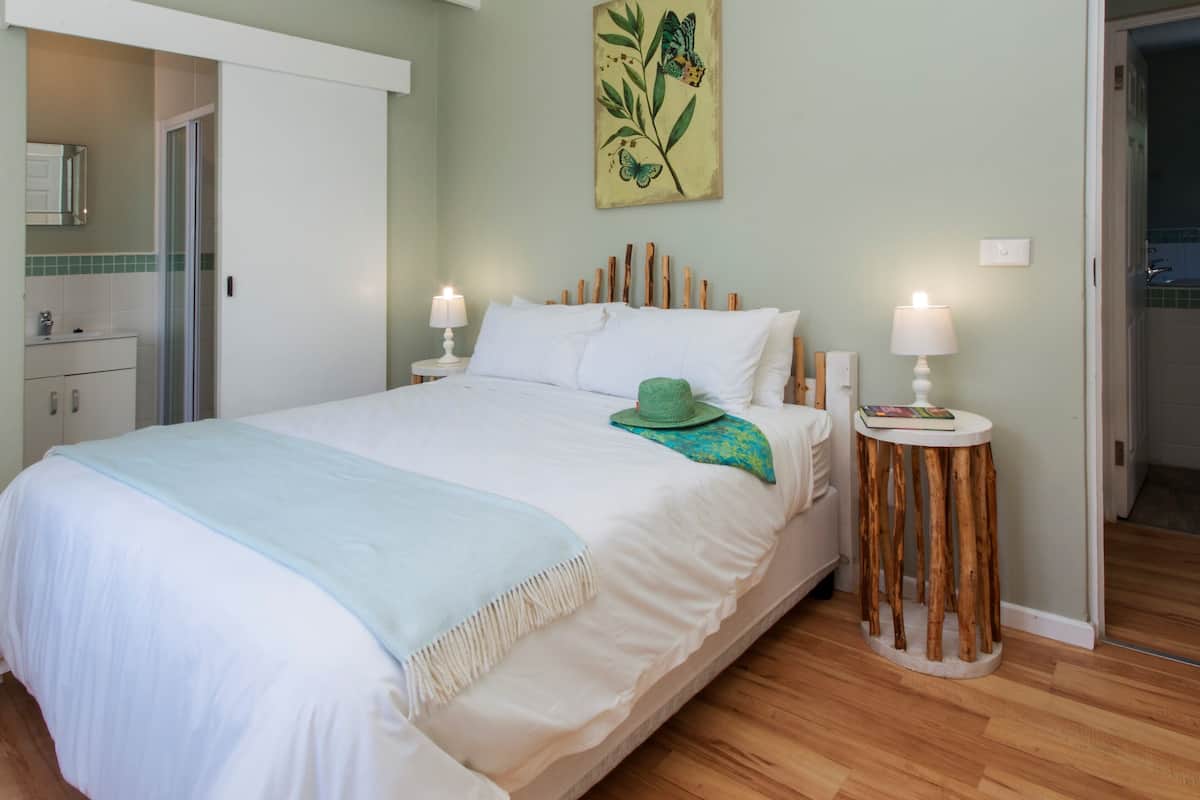 Een net opgemaakt bed met witte lakens en een groene hoed op het kussen, een houten hoofdbord, twee nachtkastjes met lampen en een aan de muur hangend schilderij van een plant.