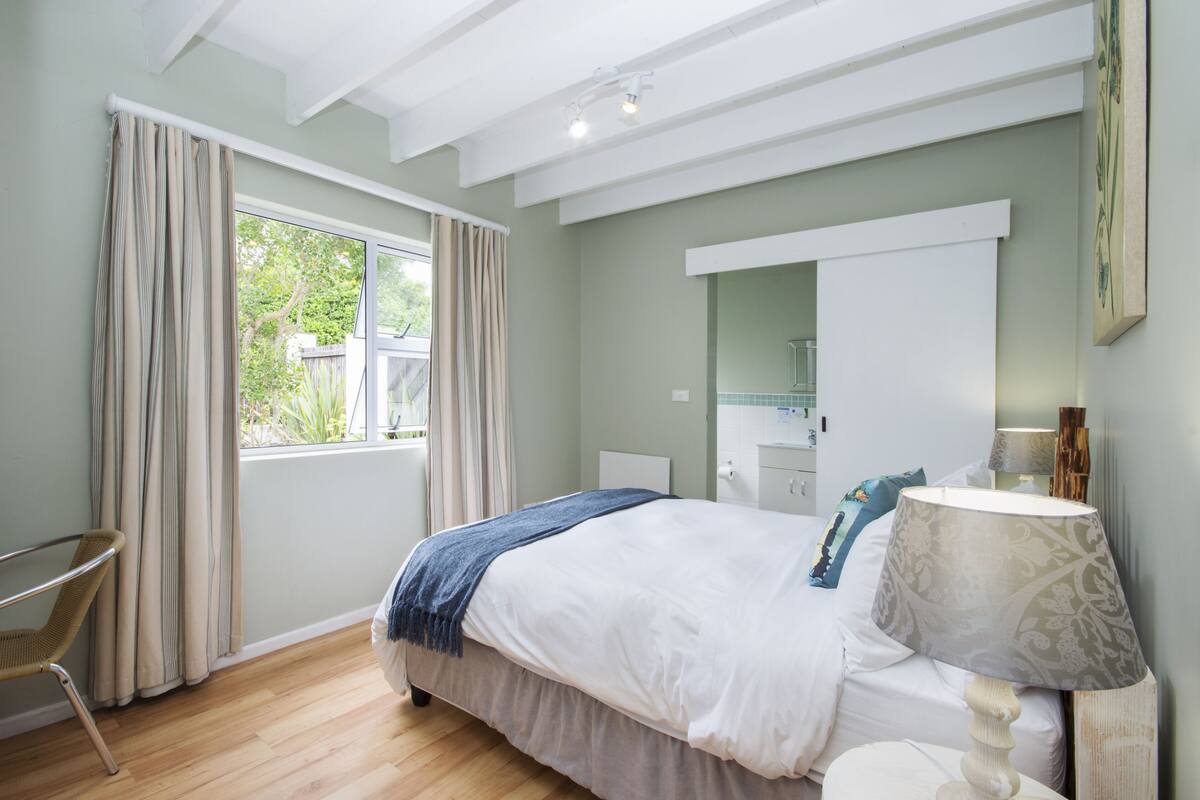 Een slaapkamer met een bed, een stoel, een nachtkastje met een lamp, een raam met gordijnen en een spiegel aan de muur.