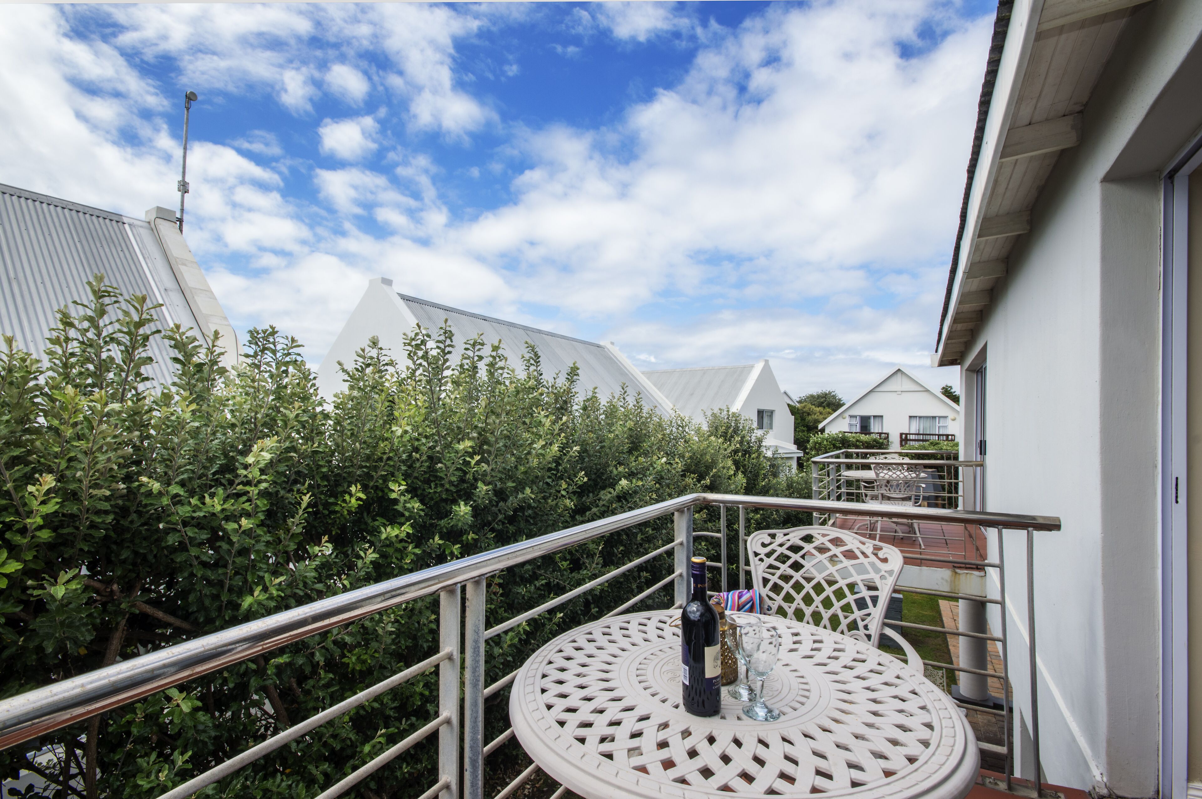 A balcony with a metal railing, a round table with a bottle and glasses, and a view of neighboring houses and greenery.