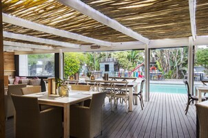 An outdoor patio area with wooden tables and wicker chairs, a pool, and a view of the surrounding greenery.