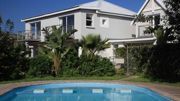 A large house with a swimming pool, surrounded by lush greenery and a clear blue sky.