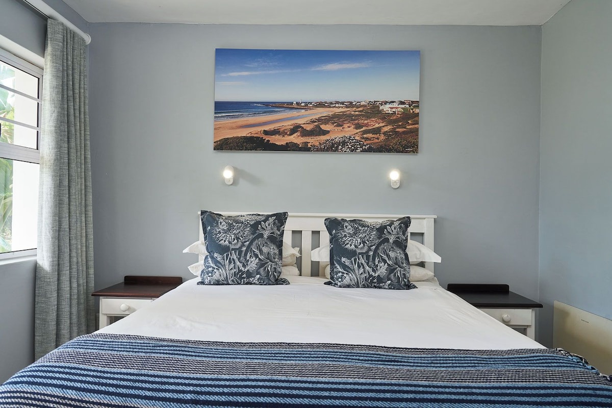 A bedroom with a bed, two pillows, a nightstand, a wall-mounted picture, and a window with curtains.